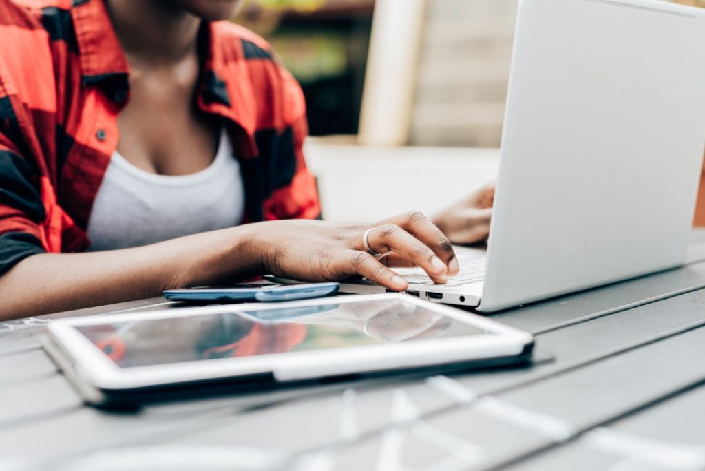 Close up on the hands of young black woman tapping the screen of a computer, surrounded by devices like smart phone and tablet - business, working, multitasking concept