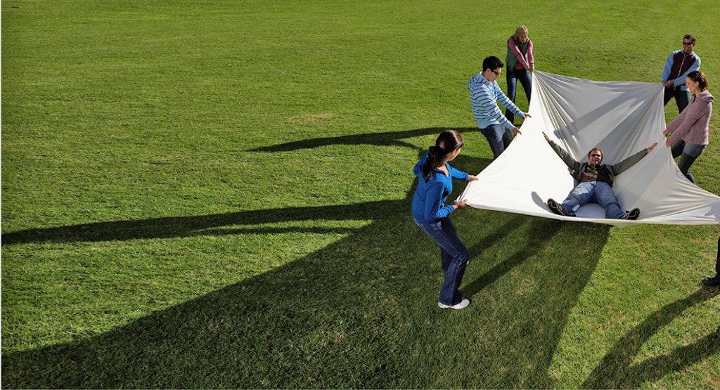 Group of people in park holding large white sheet with man lying inside